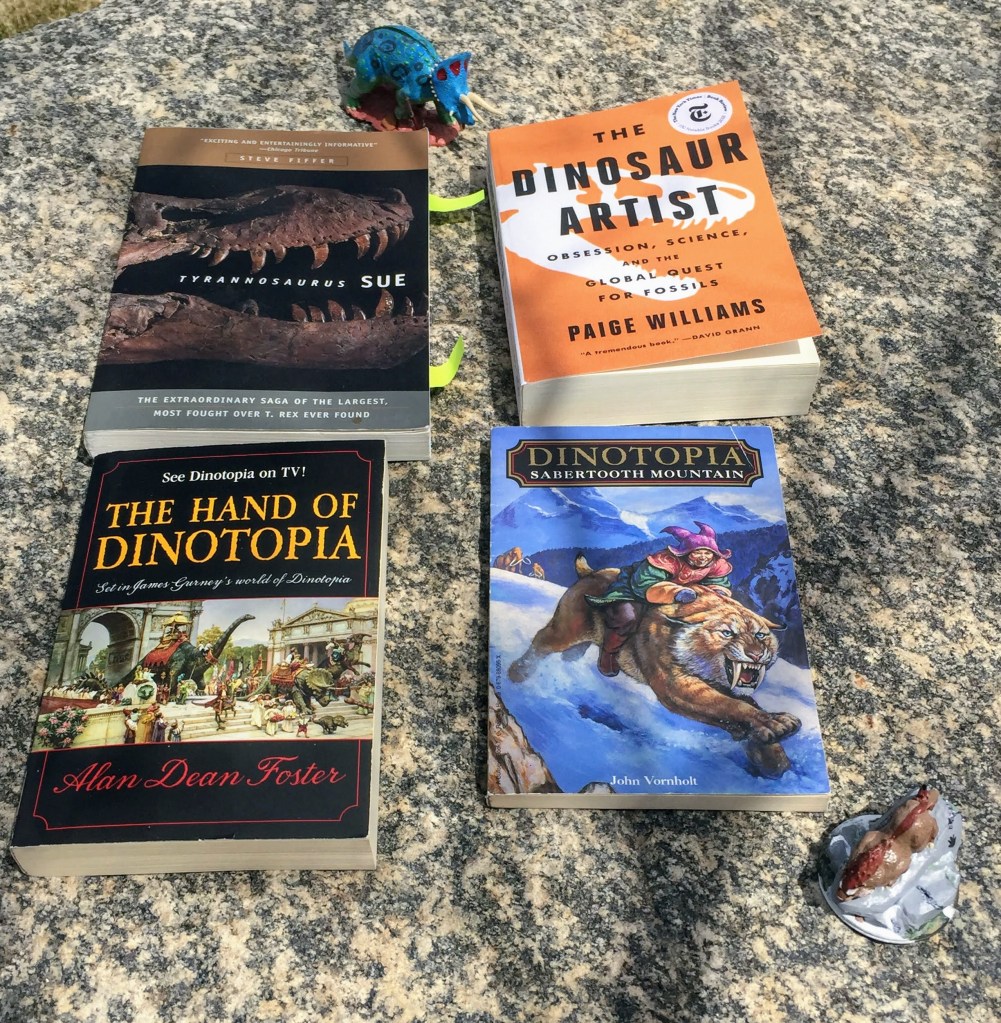Four books sitting on a grey granite rock. The top left is "Tyrannosaurus Sue," which has a black cover and a brown tyrannosaur skull on it. The Top right is "The Dinosaur Artist" which has an orange cover with a white tyrannosaur skull on it. Bottom left is "The Hand of Dinotopia" with a black cover and illustrations of a dinosaur-inhabited city on it. The bottom right book is "Sabertooth Mountain," which has a boy in a purple hat riding a sabertooth down a snow-covered mountain on its cover.