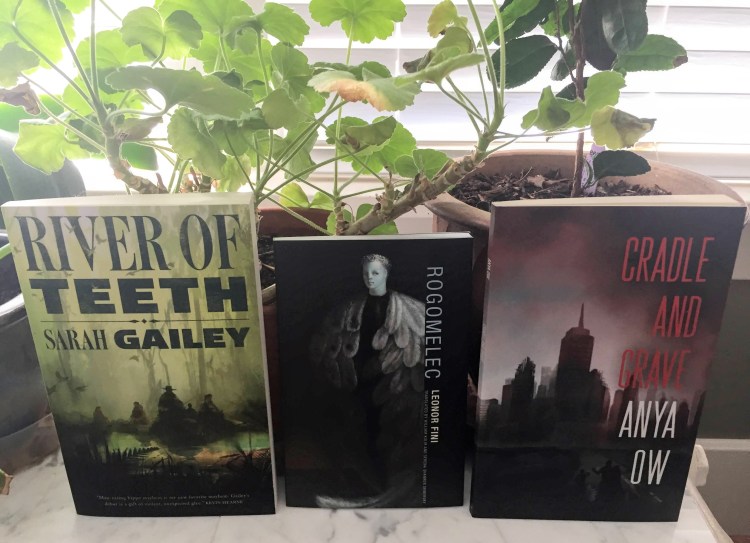 Three books propped up against two potted plants. The book on the left is River of Teeth by Sarah Gailey. It has a green cover with people riding hippos. The middle book is Rogomelec by Leonor Fini. It has a black cover with a blue winged individual on it. THe book on the right is Cradle and Grave by Anya Ow. It has a grey and red cover with a ruined city in the foreground. Green elaves rise from the pots above the books.