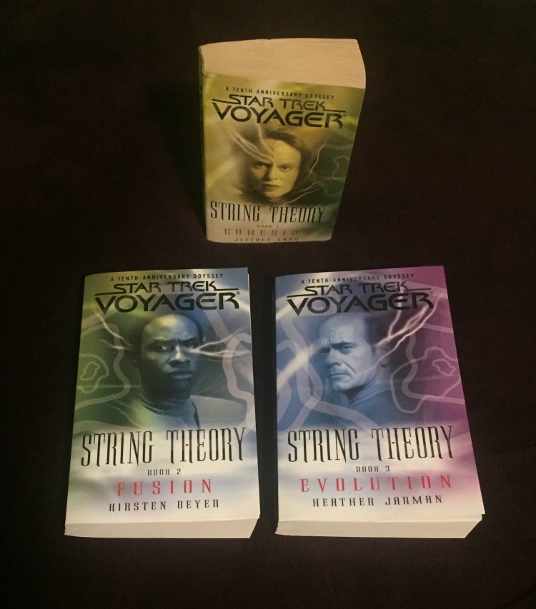 The three books from the Star Trek: Voyager String Theory trilogy sitting on a purple background. The top book has a primarily yellow cover with B'Elanna Torres on it. The other two books are sitting below the first one. The second book is green and blue with Tuvok on the cover, and the third book is blue and purple with the Doctor on the cover.