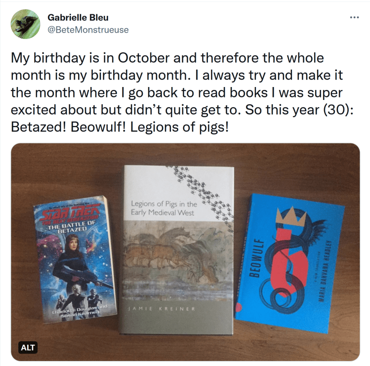 A screenshot of a tweet reading "My birthday is in October and therefore the whole month is my birthday month. I always try and make it the month where I go back to read books I was super excited about but didn’t quite get to. So this year (30): Betazed! Beowulf! Legions of pigs!" A photo is attached to the tweet. In the photo, three books are laid out side-by-side. The leftmost book is a paperback of “Battle for Betazed.” Deanna Troi is on the cover holding a gun. The center book is a large hardcover of “Legions of Pigs in the early Medieval West.” The cover has a painting of a head heard of pigs. The late book on the right is a paperback of Beowulf. The cover is blue with a lock dragon entwined around a red “b.”